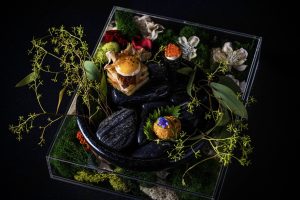 Gourmet plated dishes with seafood, edible flowers, and microgreens on black slate